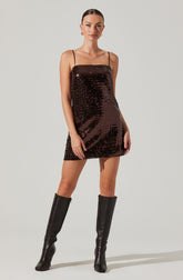 A woman is wearing a short, sleeveless dress made of shiny, dark brown sequins with thin spaghetti straps. She pairs the dress with tall black leather boots that reach just below her knees.